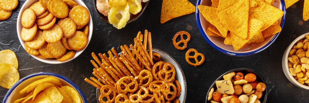 Salty snacks, party mix panorama. A variety of crispy appetizers, overhead flat lay shot. Potato and tortilla chips, crackers, popcorn etc panoramic banner
